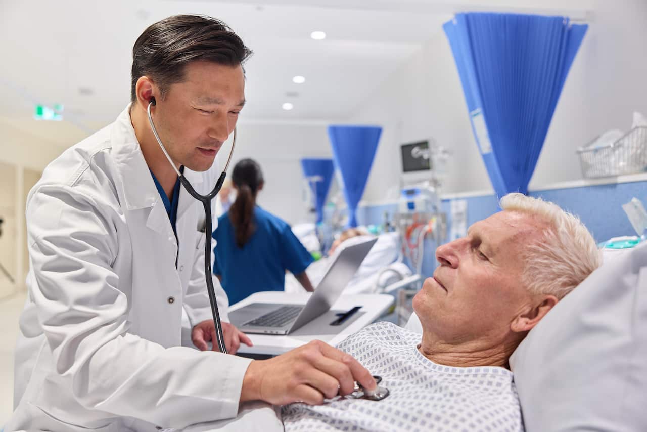 A doctor holds a stethoscope to a man's chest while he lies in a hospital bed.