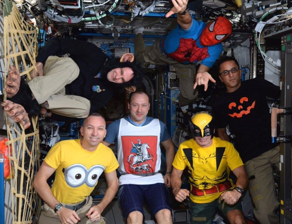A group of six astronauts wearing an assortment of costumes pose in a space station module.