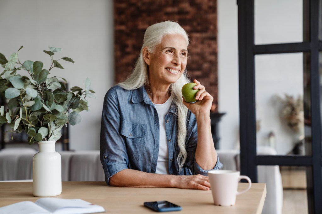 A smiling senior woman sits at a kitchen table, holding a green apple.