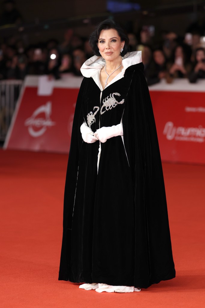 Kris Jenner on the red carpet wearing a black cape with embellished scorpion details, a white collar, and white gloves.