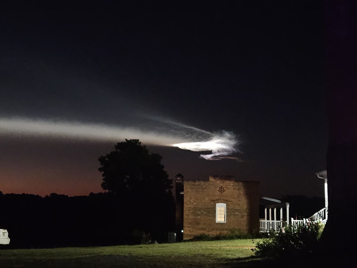 a white streak illuminated in the sky flies over a dark landscape and a brick house.