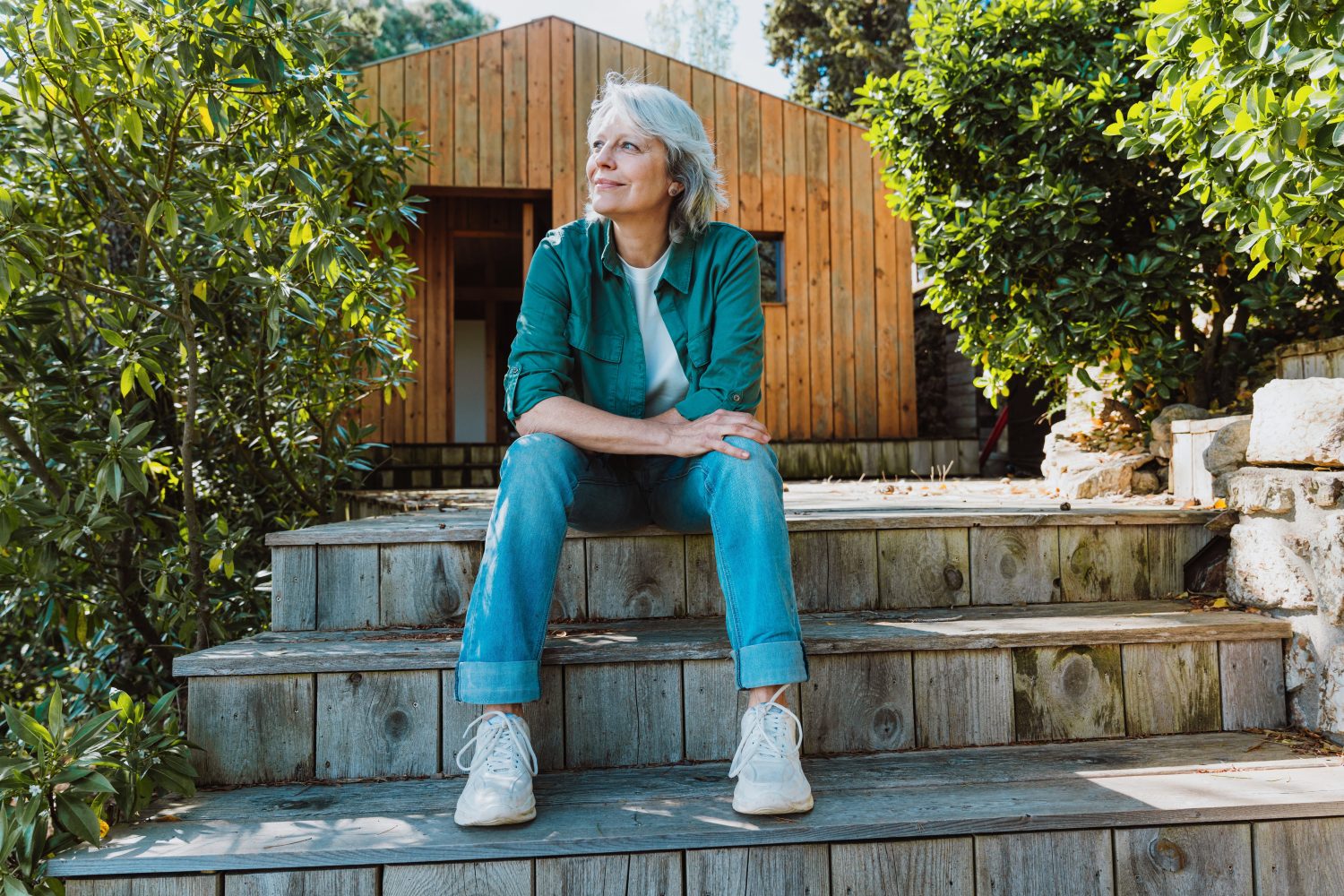 Senior woman relaxing on rustic wooden stairs, looking thoughtfully into the distance, embracing a peaceful outdoors retirement lifestyle at her simple wooden cabin surrounded by nature