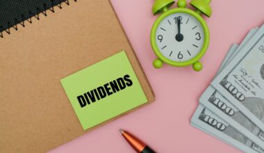 A top-down view of financial items on a soft pink surface. A brown spiral notebook with a black wire binding is positioned on the left. A bright green sticky note with the word 'DIVIDENDS' in bold black text is adhered to the notebook. To the right, a small, vibrant green analog alarm clock with a white face shows the hands close to twelve o'clock. Three stacks of US $100 bills are fanned out in the bottom right corner, and a black pen with an orange grip lies in the lower center.