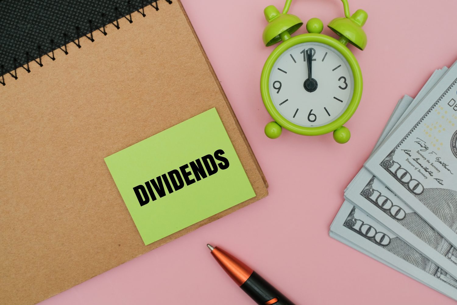 A top-down view of financial items on a soft pink surface. A brown spiral notebook with a black wire binding is positioned on the left. A bright green sticky note with the word 'DIVIDENDS' in bold black text is adhered to the notebook. To the right, a small, vibrant green analog alarm clock with a white face shows the hands close to twelve o'clock. Three stacks of US $100 bills are fanned out in the bottom right corner, and a black pen with an orange grip lies in the lower center.