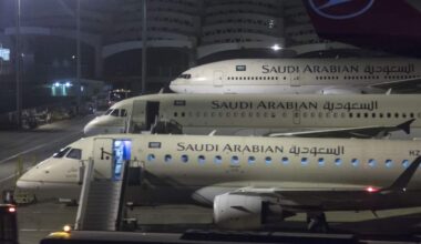 Three Saudi Arabian Airlines aircraft are parked on an airport apron at night, viewed through a terminal window. A regional jet is in the foreground with boarding stairs attached and cabin lights visible through the open door. Behind it, two larger Saudi Arabian Airlines aircraft are lined up nose-to-tail. Airport service vehicles, floodlights and terminal infrastructure are visible around the aircraft. In the upper-right corner, part of a Turkish Airlines aircraft tail with the airline logo can be seen. The scene is illuminated by bright airport lighting against a dark night sky.