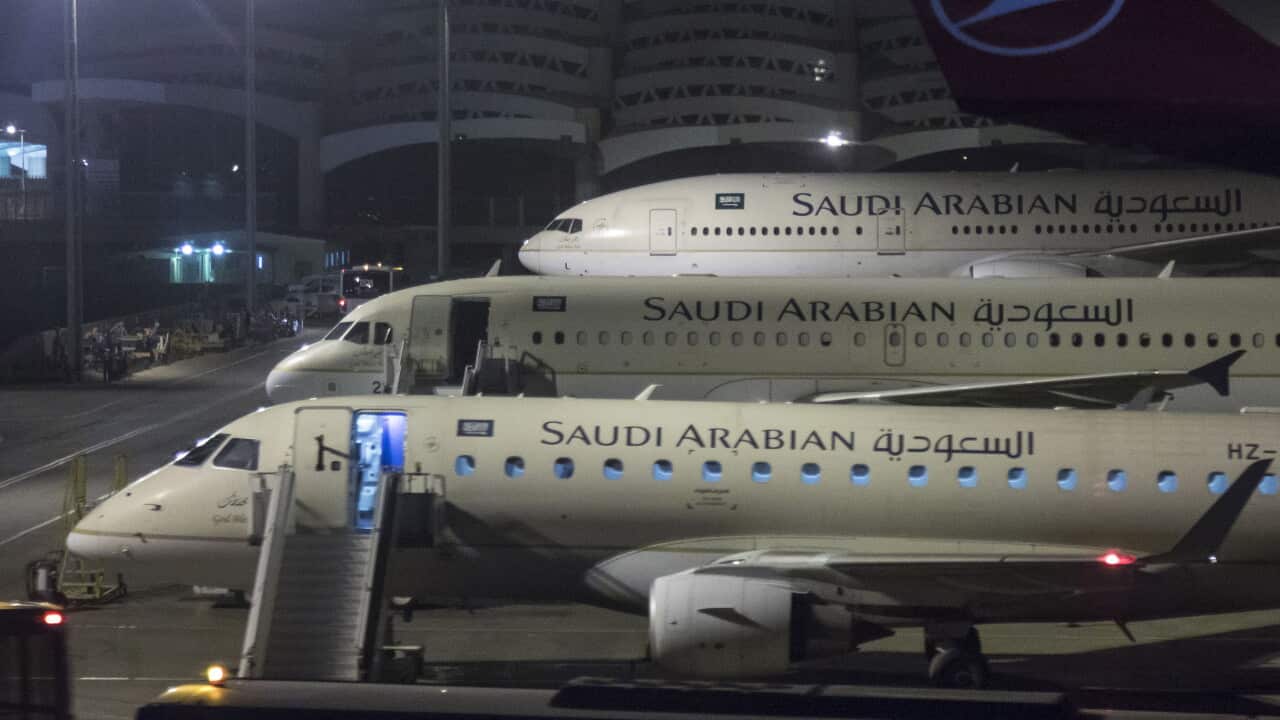 Three Saudi Arabian Airlines aircraft are parked on an airport apron at night, viewed through a terminal window. A regional jet is in the foreground with boarding stairs attached and cabin lights visible through the open door. Behind it, two larger Saudi Arabian Airlines aircraft are lined up nose-to-tail. Airport service vehicles, floodlights and terminal infrastructure are visible around the aircraft. In the upper-right corner, part of a Turkish Airlines aircraft tail with the airline logo can be seen. The scene is illuminated by bright airport lighting against a dark night sky.