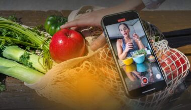A reusable mesh grocery bag filled with fresh produce, including celery, leeks, apples and a green tomato, sits on a wooden table. A hand reaches toward a red apple while a smartphone resting on the produce displays a video recording of a person preparing food in a kitchen, suggesting food content creation or online cooking advice.