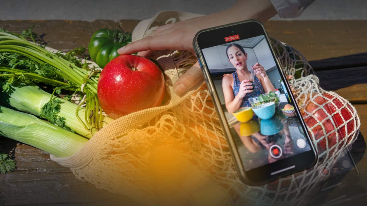 A reusable mesh grocery bag filled with fresh produce, including celery, leeks, apples and a green tomato, sits on a wooden table. A hand reaches toward a red apple while a smartphone resting on the produce displays a video recording of a person preparing food in a kitchen, suggesting food content creation or online cooking advice.