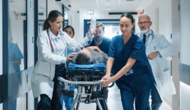 Healthcare staff rush a patient on a hospital stretcher down a brightly lit corridor, with multiple clinicians accompanying the stretcher and one staff member holding an IV bag overhead during an apparent emergency transfer.