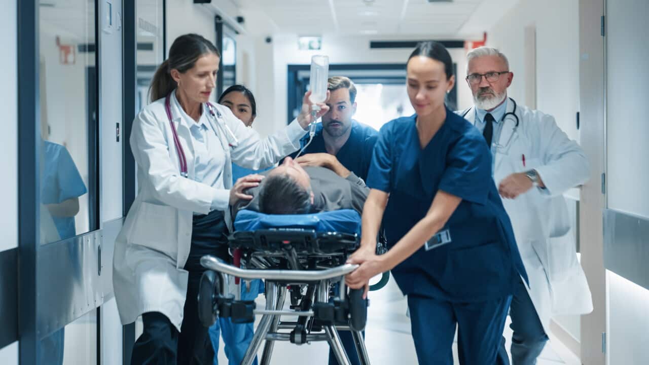 Healthcare staff rush a patient on a hospital stretcher down a brightly lit corridor, with multiple clinicians accompanying the stretcher and one staff member holding an IV bag overhead during an apparent emergency transfer.
