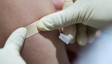 A close-up image of a healthcare worker wearing disposable gloves applying a small adhesive bandage to a person's upper arm at the site of an injection. The image focuses on the bandage being pressed onto the skin, with the background softly blurred.