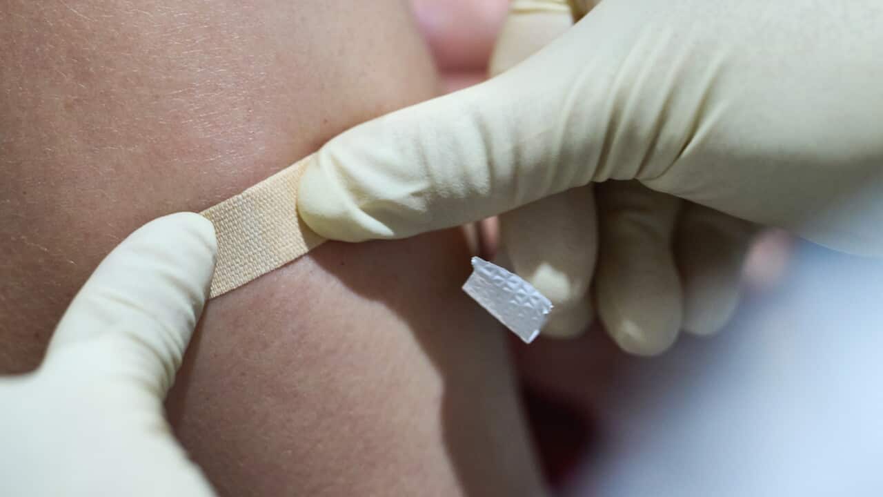 A close-up image of a healthcare worker wearing disposable gloves applying a small adhesive bandage to a person's upper arm at the site of an injection. The image focuses on the bandage being pressed onto the skin, with the background softly blurred.