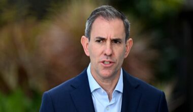 A middle aged man with greying hair, dressed in a dark blue blazer with blight blue button-down shirt, is speaking. The background behind him is blurred.