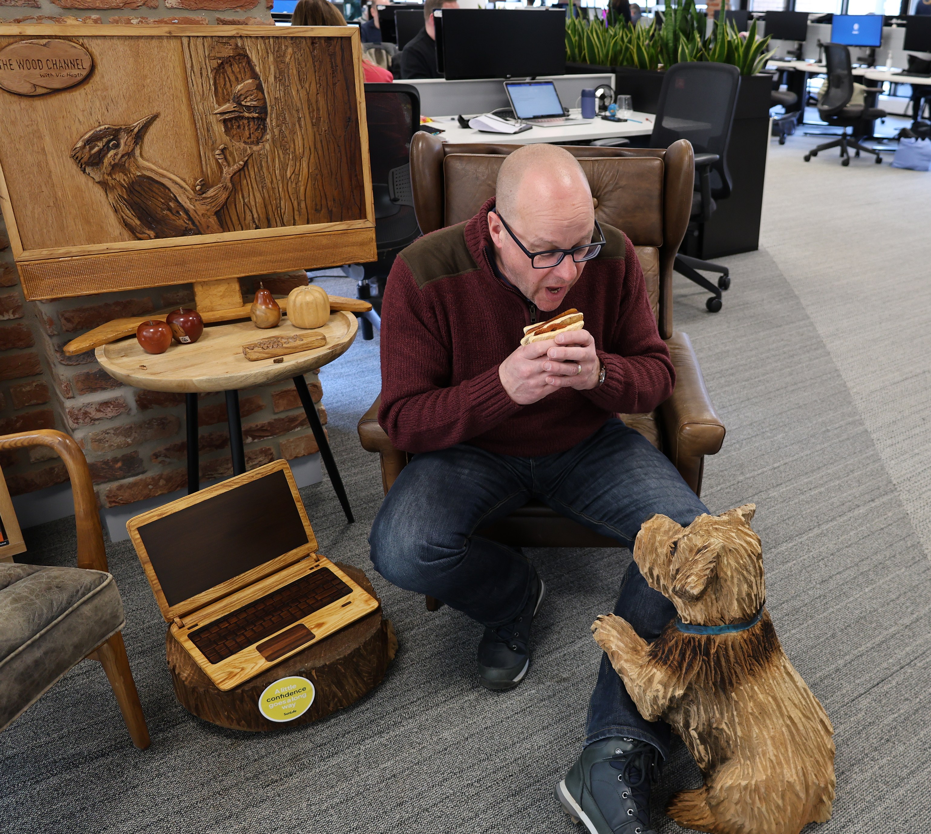 Man eating a sandwich while sitting in a chair with a carved wooden dog at his feet.  Wooden laptop and woodpecker carving also visible.