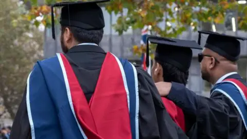Getty Images Three men wearing black, red and blue graduation gowns and black caps. They have their backs to the camera, with their arms round each other, stood under a tree. They all have black hair and one is wearing sunglasses. 