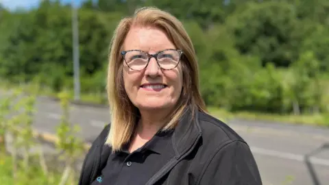 Karen Jones, who runs GoodLives charity in Cumbria, is pictured smiling. She has brown hair with blonde highlights and is wearing glasses which have dark frames. She is wearing a black work uniform which displays the charity's logo.