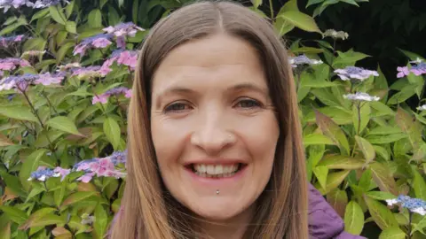 Jo Boylan Jo Boylan, a woman with long, straight, fair hair, smiles into the camera.  She is wearing a purple puffed jacket and has a small silver piercing below her lip.  She is standing in front of a shrub with purple and pink flowers.
