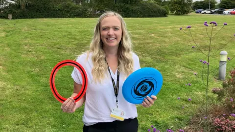 A smiling blonde woman stands on a grass bank holding two toy flying rings.