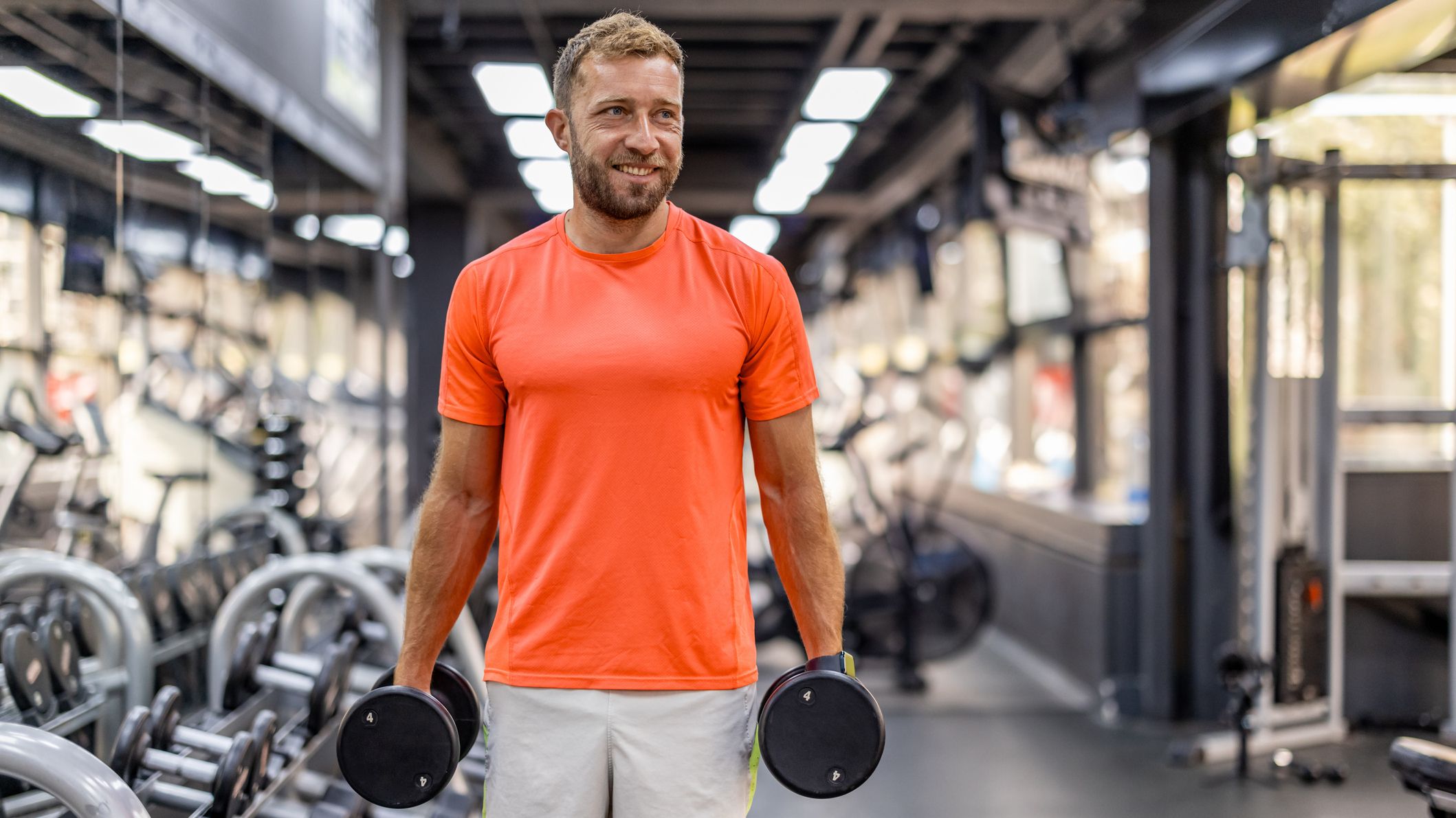 Man in gym holding dumbbells by his sides. He is wearing grey shorts and a bright orange T-shirt