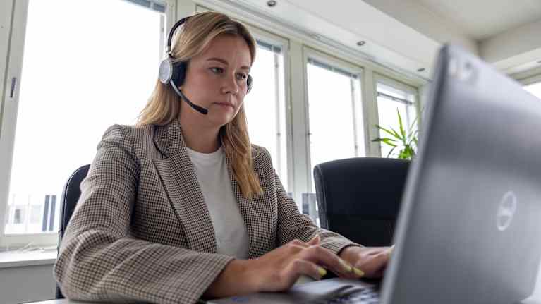 A woman sits in an office with a headset on and is typing on a laptop.