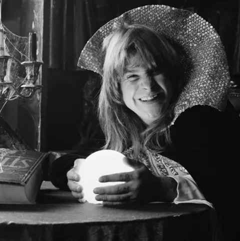 Getty Images Ozzy Osbourne holds a crystal ball while dressed in a wizard's robes. On the table in front of him is a book of spells and a candleabra encrusted with spiders' webs.