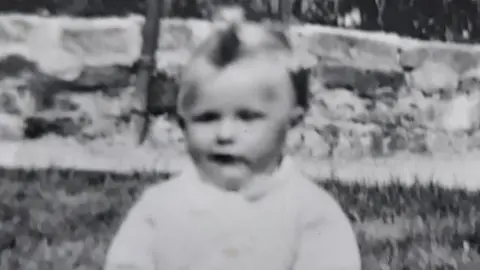 Family handout An old black and white photograph of Susan (not her real name) as a baby in the 1950s. She is wearing a white jumper and sitting on grass in front of a low brick wall.