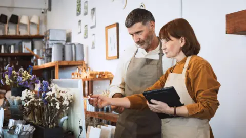 Getty Images Two middle aged people in aprons and autumnal clothing in a florists. They are looking at a screen.