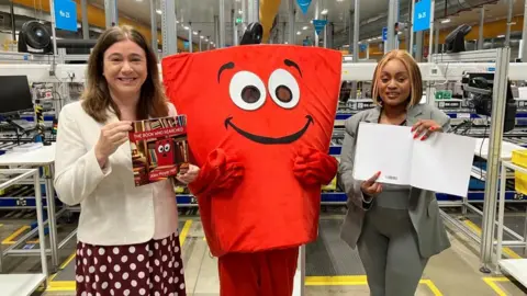 Sam Read/BBC Alex Mayer, holding a book, wears a white jacket and red and white spotted dress. There is a red cartoon character in the middle and Sally Kimondo, wearing grey leggings and a grey jacket, is next to it. 