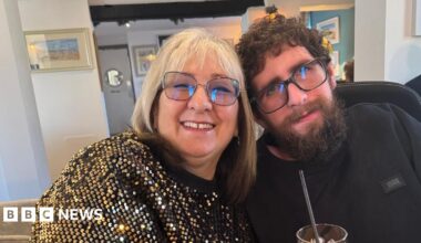Kate Bingham smiles at the camera sat next to her son Timothy - she has silver hair, glasses and wears a gold sparkly top, he is wearing all black and has glasses and a long, dark beard. Kate holds a drink with a straw in it.