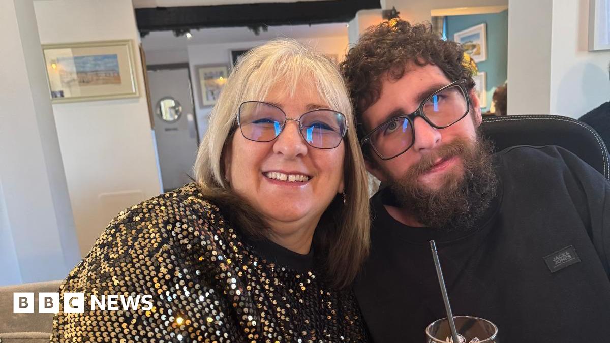 Kate Bingham smiles at the camera sat next to her son Timothy - she has silver hair, glasses and wears a gold sparkly top, he is wearing all black and has glasses and a long, dark beard. Kate holds a drink with a straw in it.