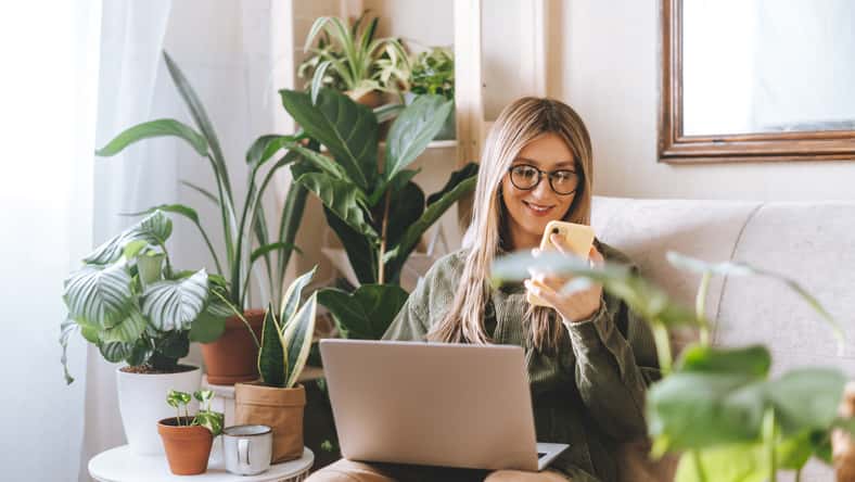 Freelance woman in glasses with mobile phone typing at laptop and working from home office. Happy girl sitting on couch in living room with plants. Distance learning online education and work.