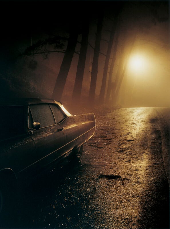 A vintage car is parked beside a wet, empty road at night, illuminated by a hazy streetlamp in the fog, with tall, shadowy trees lining the roadside.