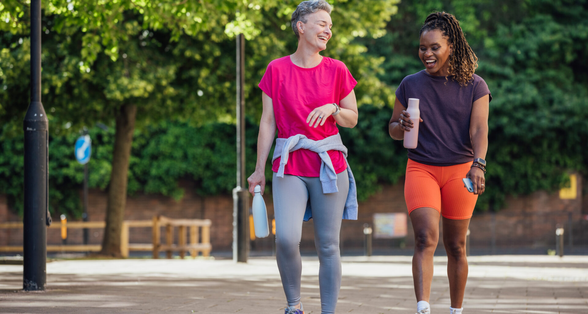 Two women walk along a paved path outside, laughing and holding water bottles. Behind them we see leafy trees and lamposts.