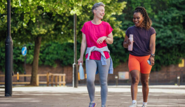 Two women walk along a paved path outside, laughing and holding water bottles. Behind them we see leafy trees and lamposts.