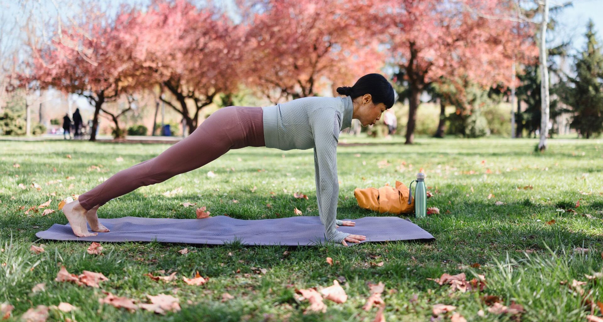 Woman practising shoulder taps in park on grass, planking on yoga mat wearing activewear in the sunshine
