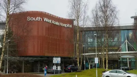Pacemaker A general view of South West Acute Hospital. The building is modern and has a copper-clad and glass front. A sign with the name of the hospital is on the top of the copper cladding in white letters. A silver car is passing by in the foreground, while bare trees without leaves are also visible.