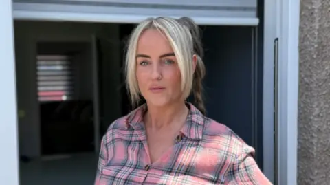 BBC A woman with blonde hair tied up in a ponytail is wearing a pink and grey checked shirt