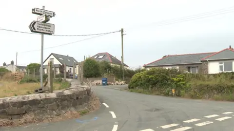 BBC A wide image of the junction into Llangwnnadl village. On the left there are road signs attached to a pole. On the right of the image are houses. 