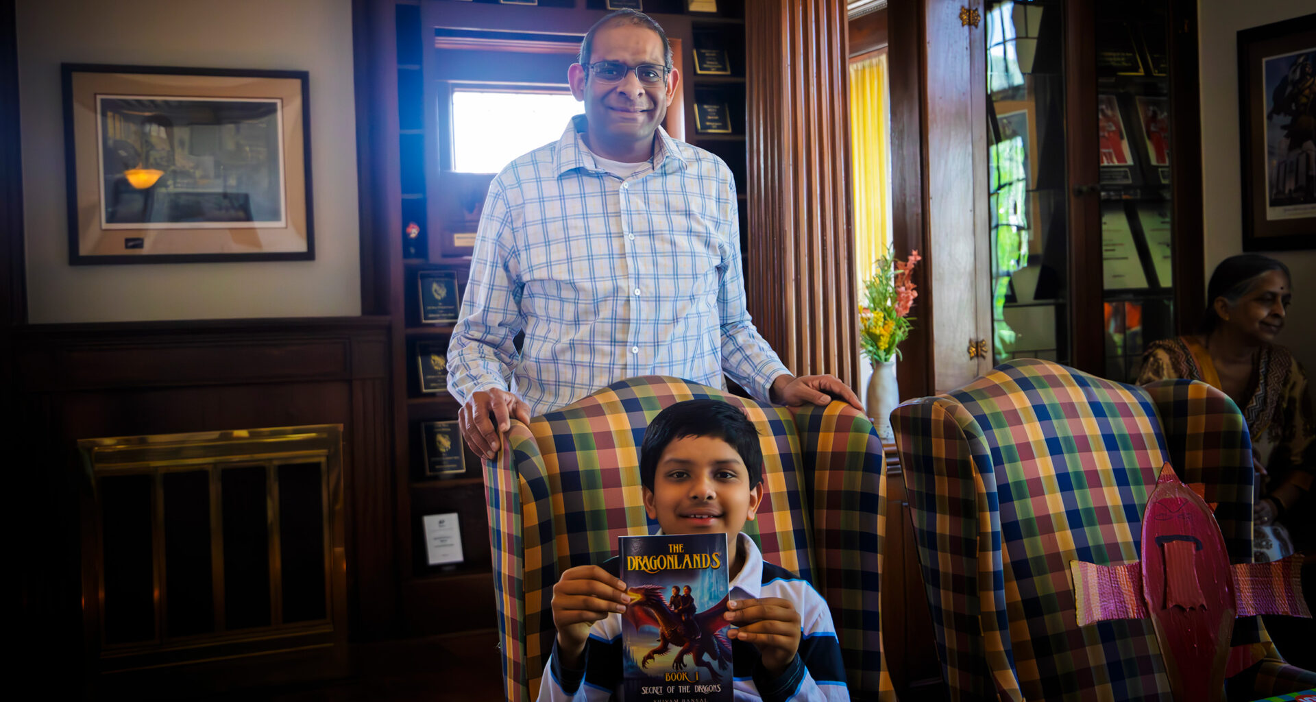 A proud father stands behind his 11-year old son. The son holds his original novel, a paperback book called "Secret of the Dragons."