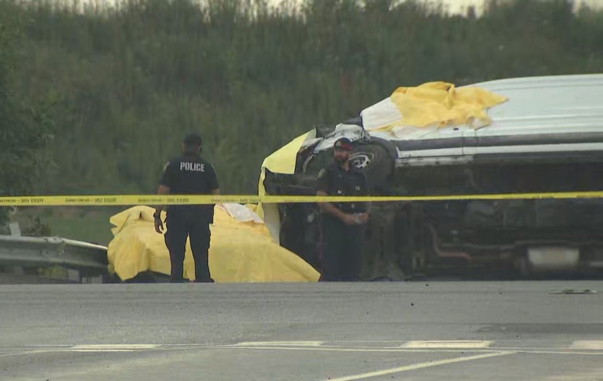 2 police officers stand by a flipped white van on a highway on a grey day. The scene is taped off