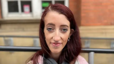 BBC News A girl with red hair and facial piercings looking at the camera, with a pink jumper on and head phones on 