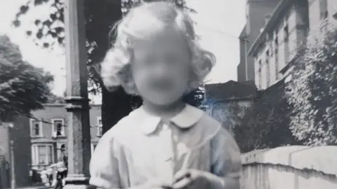 Family handout Black and white photograph of Susan (not her real name) taken in the 1950s or 1960s. She looks around seven years old and standing in a street with terraced housing behind her and a wall to her left. She has long blonde hair and is wearing a dress.