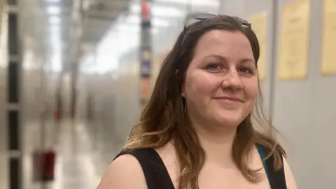 Mousumi Bakshi/BBC Image of library and archive assistant Nicki Carter wearing a black vest standing in front of some book shelves.