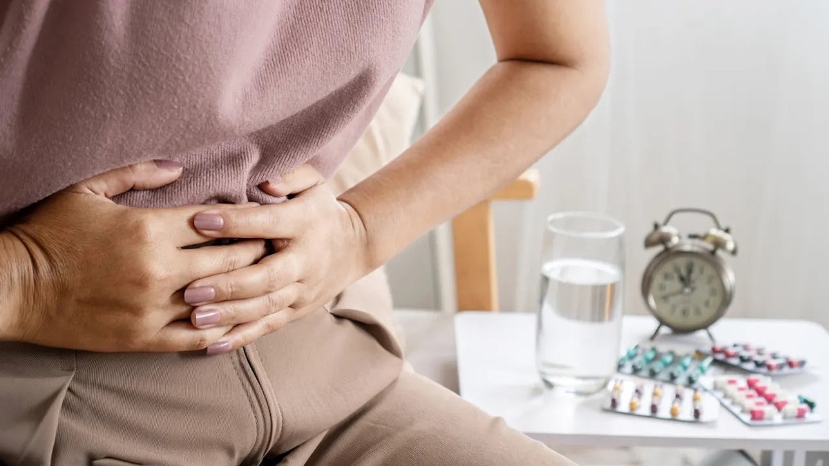 Woman clutching her stomach, apparently suffering from digestive issues, with supplements seen behind her.