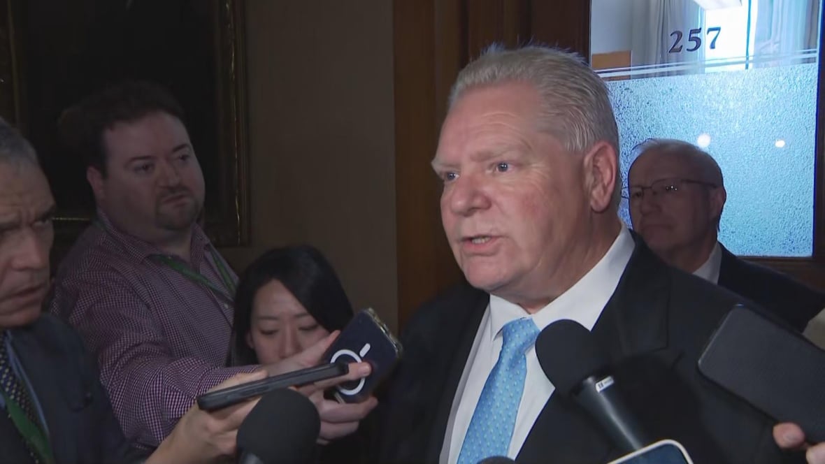 Doug Ford surrounded by reporters and speaking to a microphone in a dark-lit room 