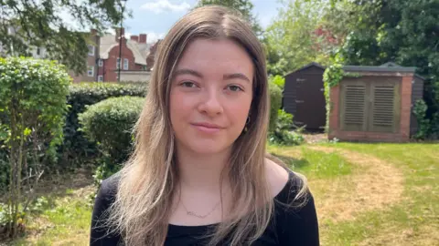 Alaw Simpson who has long blonde hair and is wearing a black t-shirt. She is stood in a garden with green bushes and trees and wooden sheds behind her.