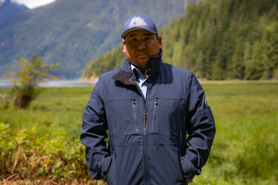 An Indigenous man stands outside, with water and a tree-covered mountain behind him.