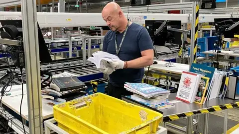 Sam Read/BBC A worker in an Amazon print factory, looking at a book. There are lots of books around him. including a big yellow box. Machinery is behind him too. 