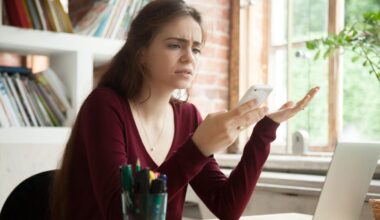 Frustrated young woman looking at her phone while sitting at her home desk where her laptop is open.
