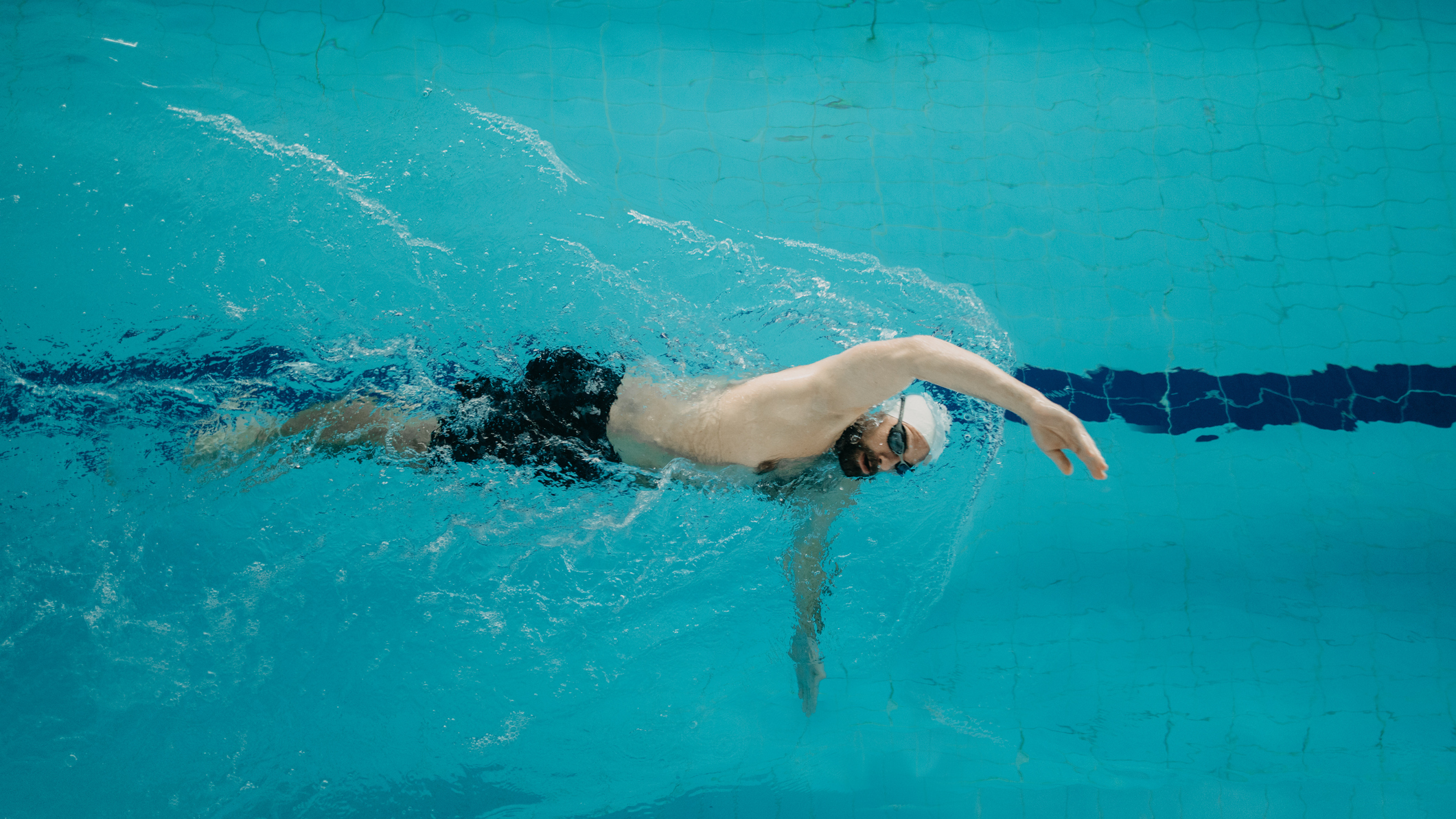 Man swimming front crawl in a pool, wearing a white swim cap, goggles and black trunks